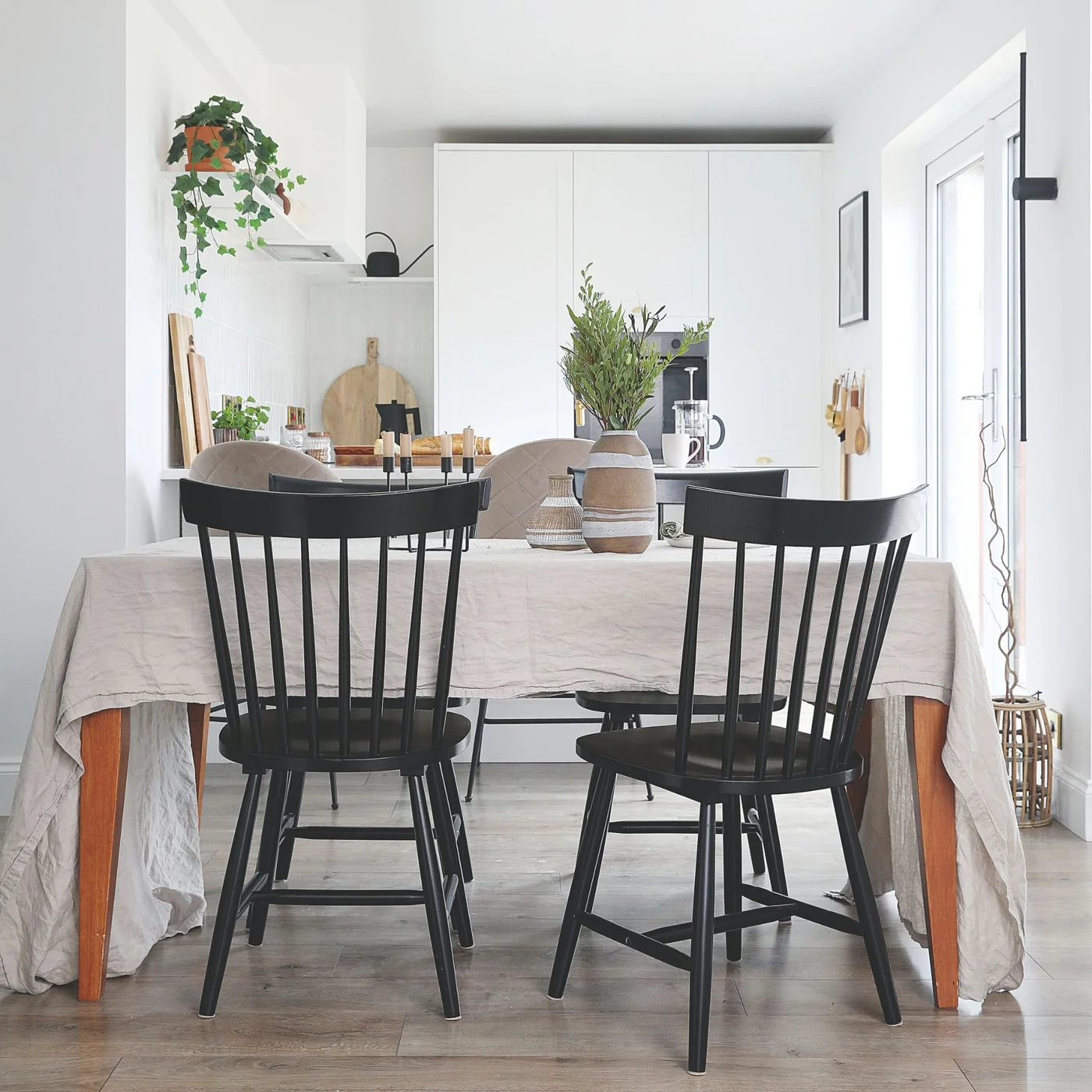 Black spindle chairs stand around a cloth-covered wooden table beside a white kitchen and a small pale vase