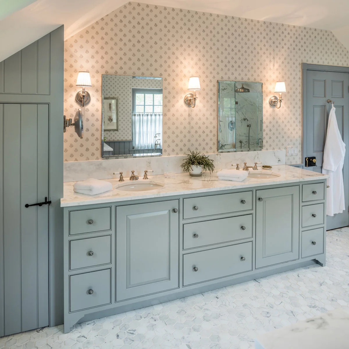 A long muted blue-gray double vanity sits beneath small patterned wallpaper, two rectangular mirrors and shaded wall lights.