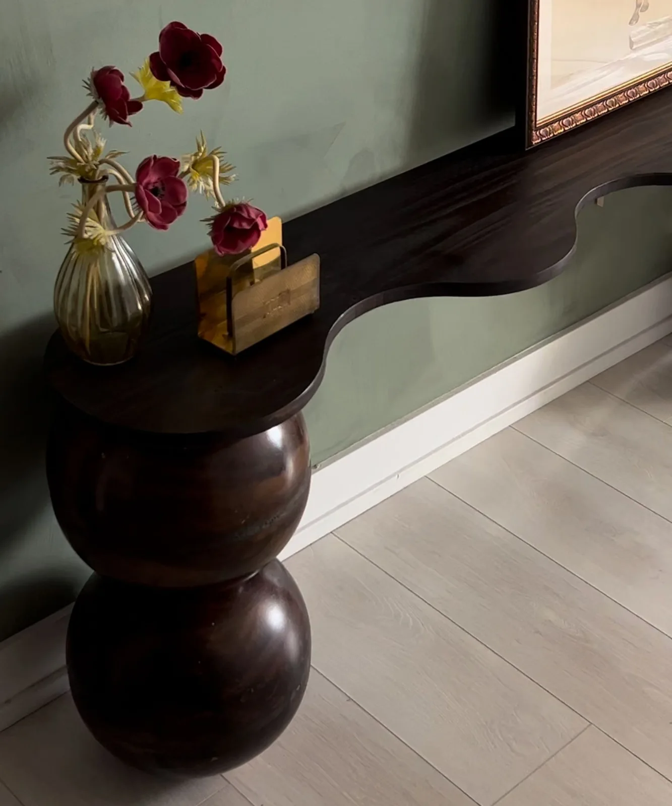 Dark narrow console with a wavy top and large rounded legs against a green hallway wall