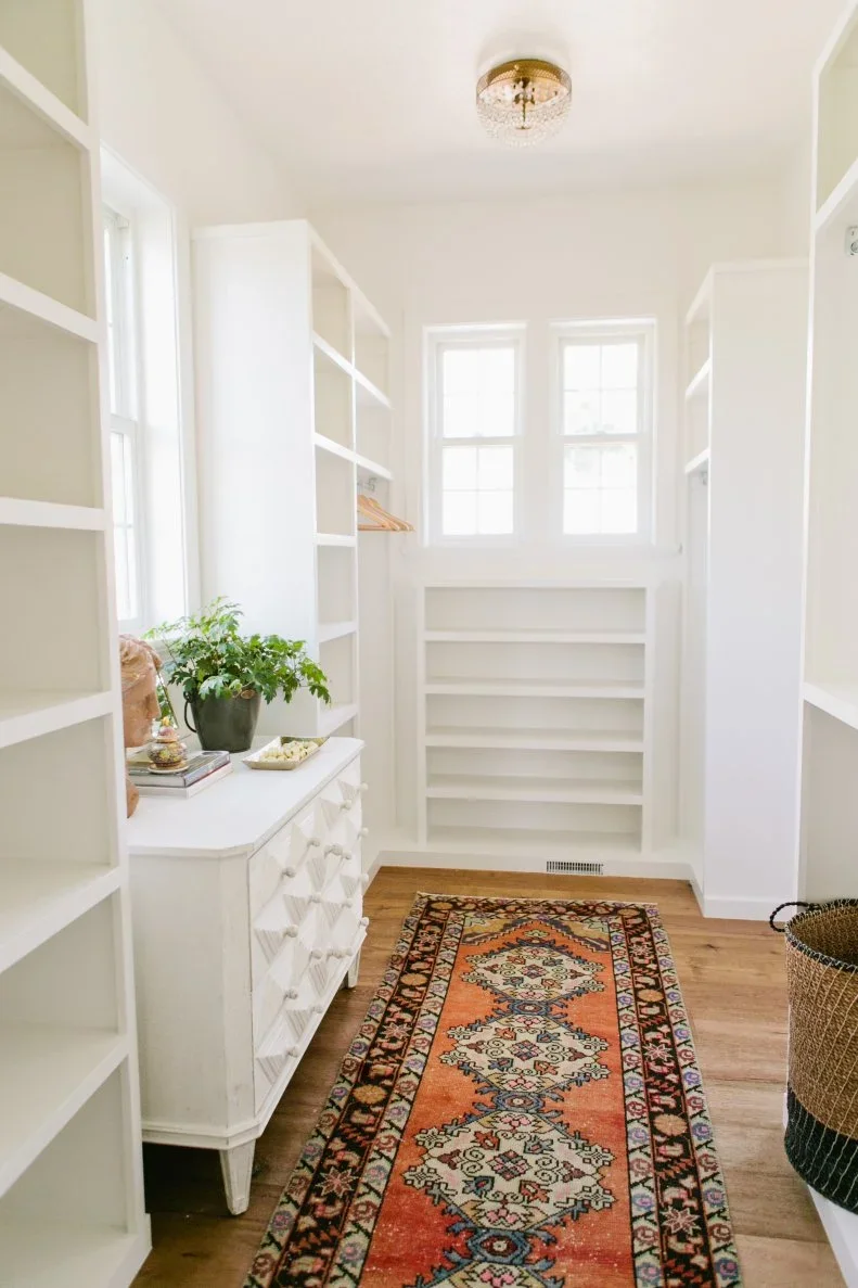 A narrow white closet has open shelves, a small drawer chest, windows and a patterned runner.