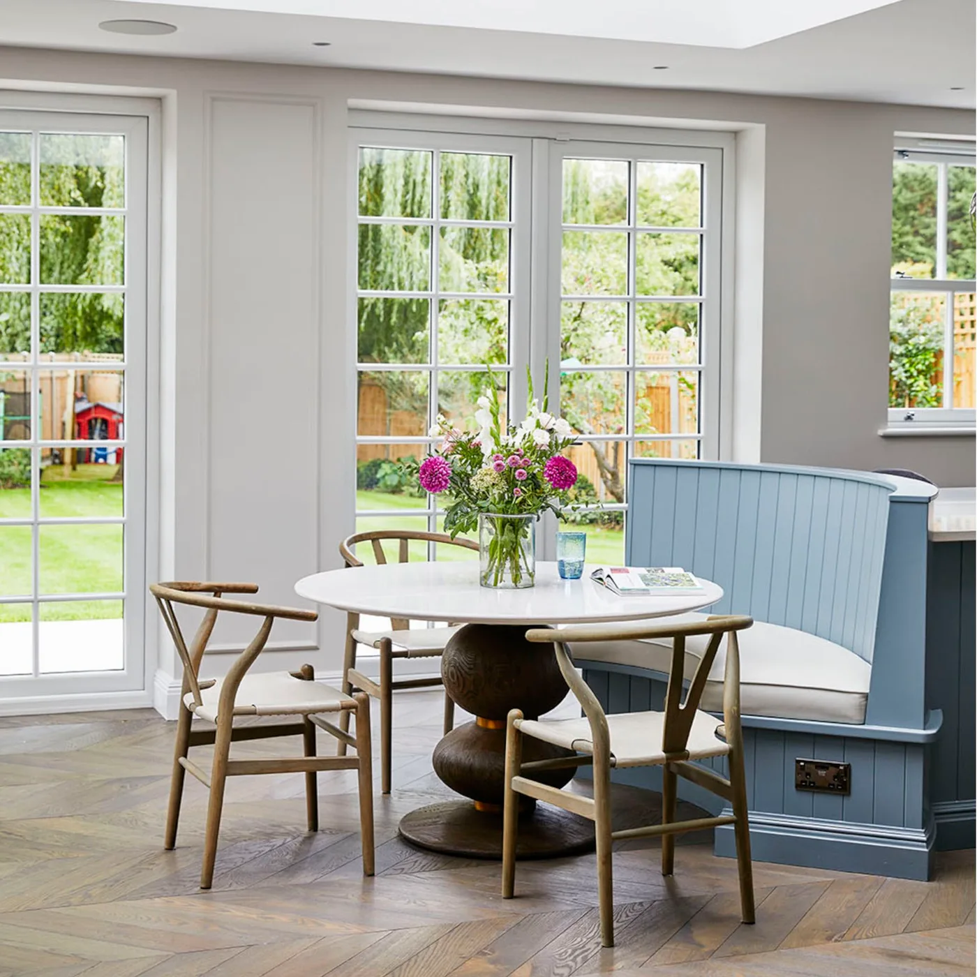 Dining nook with a curved blue banquette, pale cushions, round white pedestal table, wooden chairs and glazed doors.