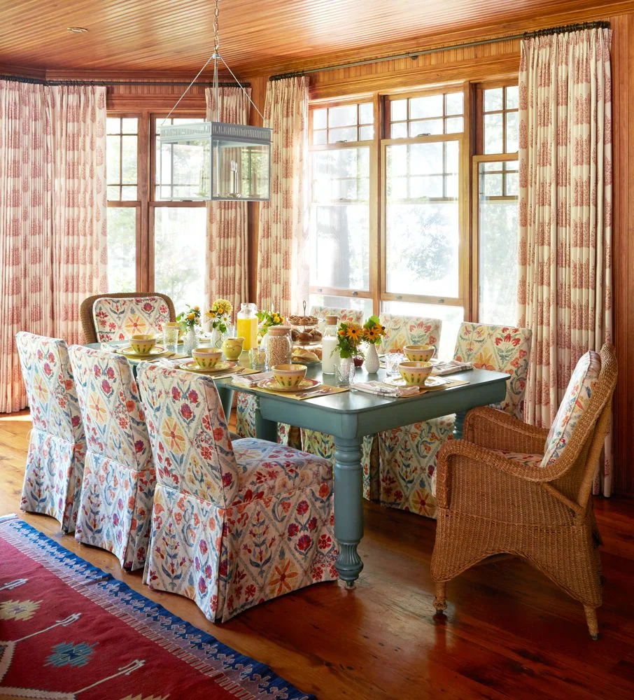 Floral-covered chairs surround a muted teal table beneath a glazed lantern beside patterned curtains and a red-and-blue rug