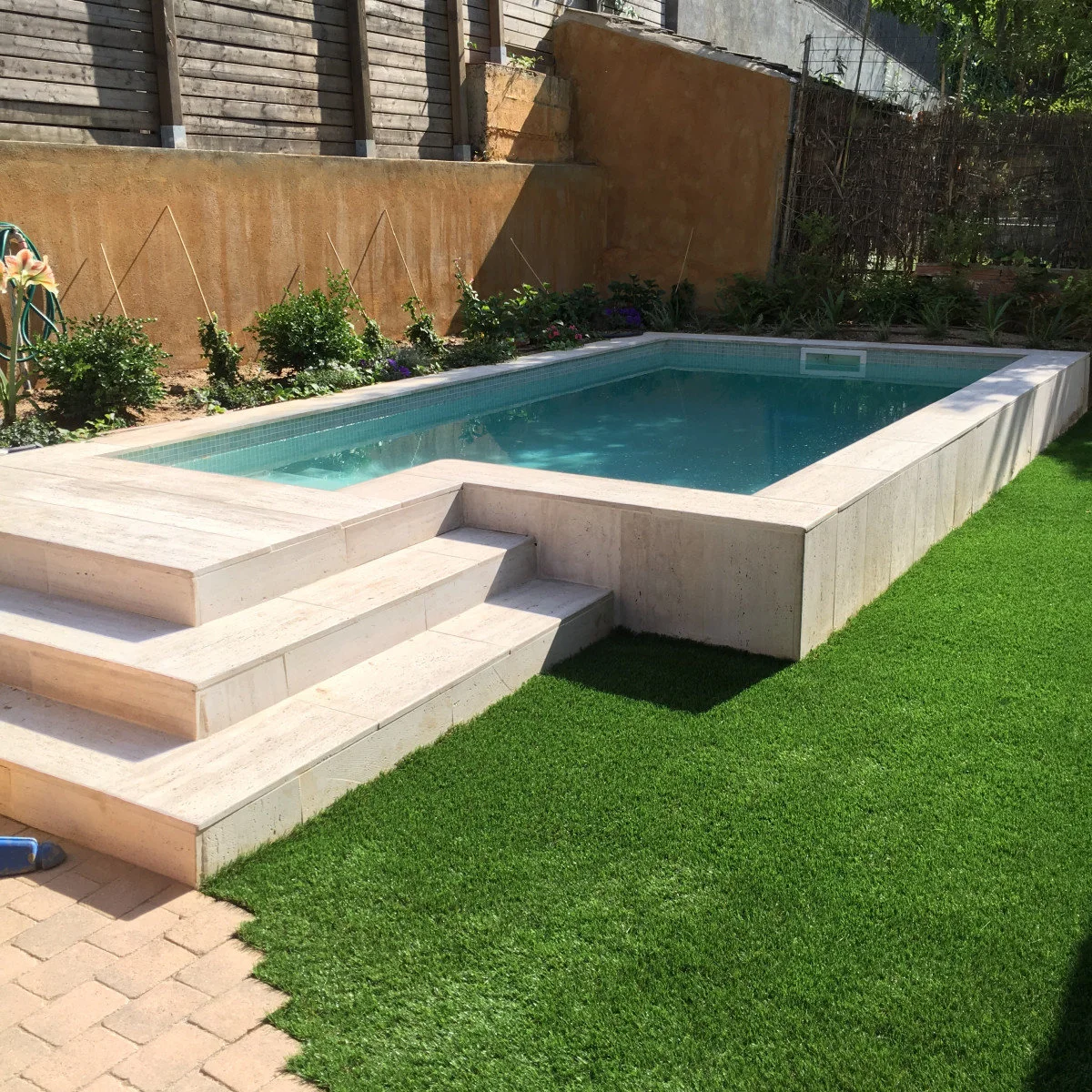 Small rectangular raised plunge pool with pale sides, broad steps and a planted border against a warm-toned wall.