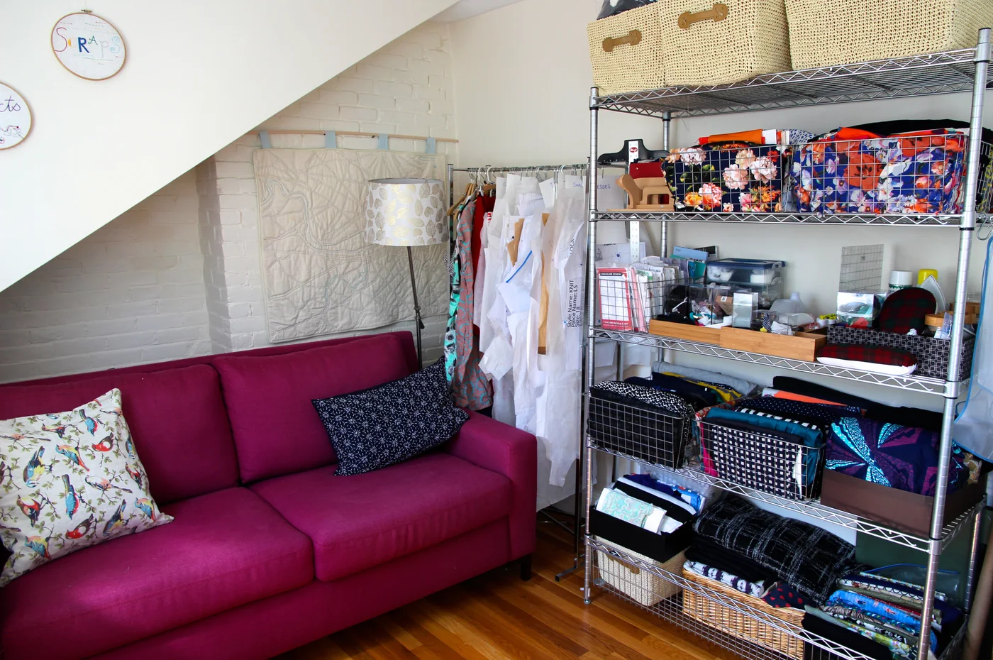 A fuchsia sofa bed sits beside metal shelving filled with fabric baskets, folded materials and sewing supplies in a room with a sloping ceiling.