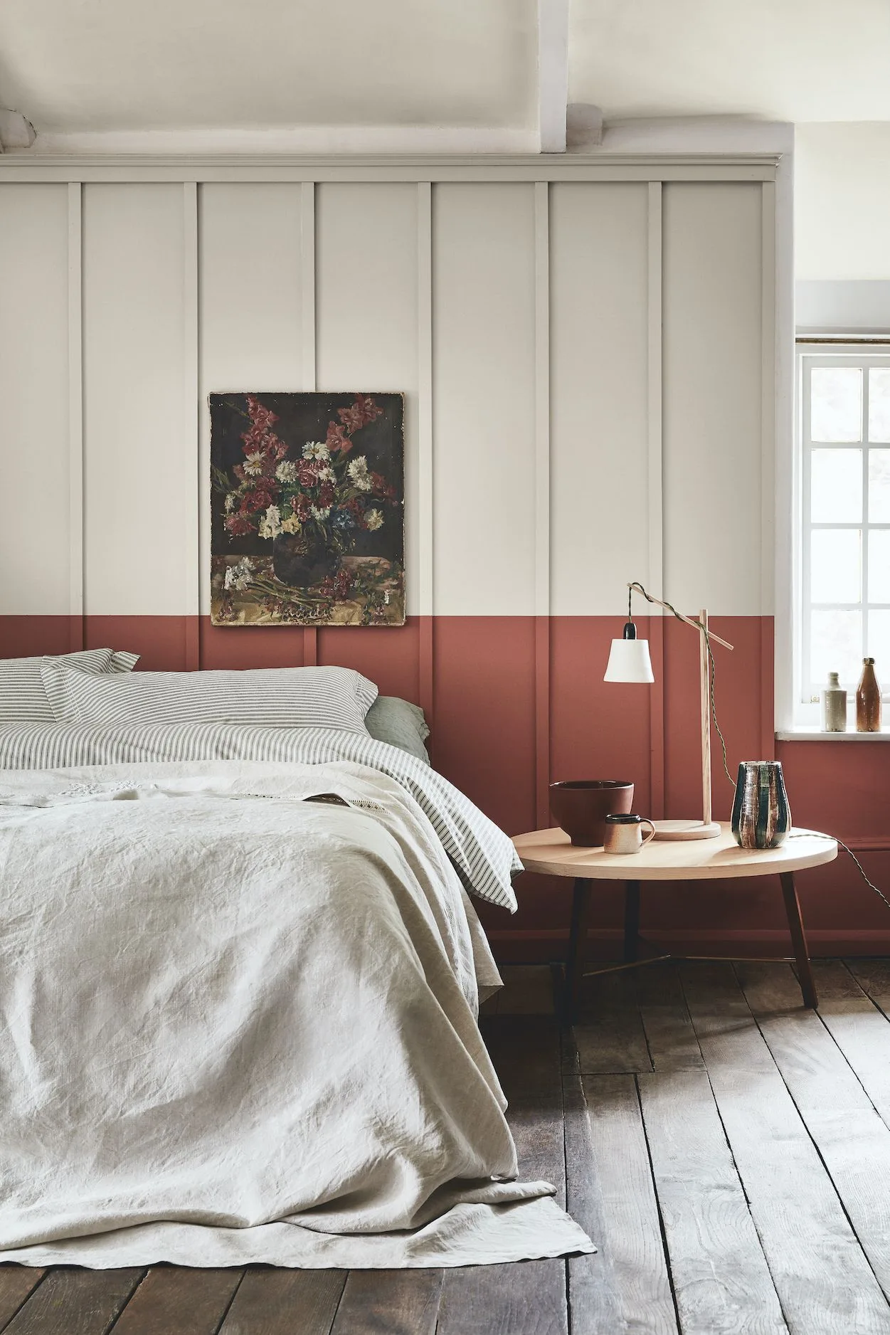 A bedroom paneled wall is painted pale above a rust-red band beside light bedding and a round timber table.