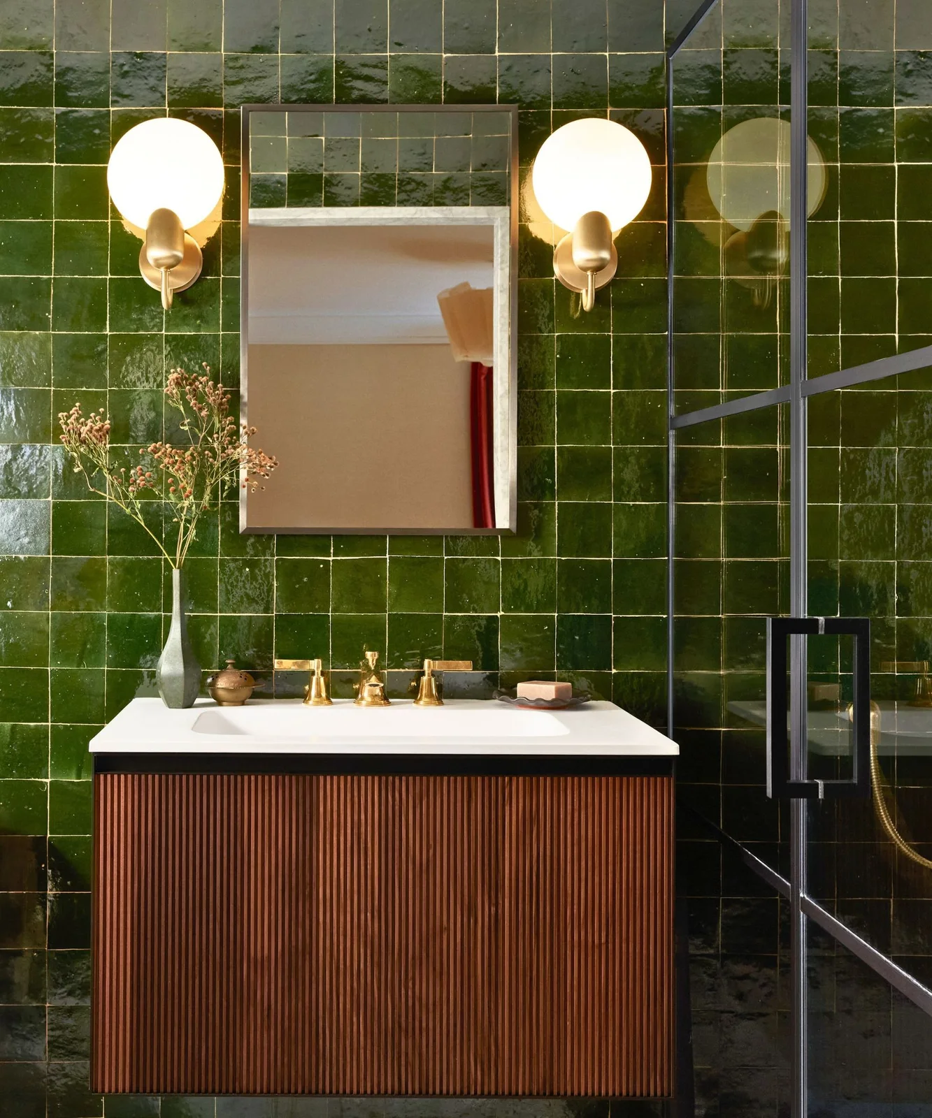 Varied olive square tiles surround a floating reeded wood vanity with a white counter, brass fixtures and globe lights.