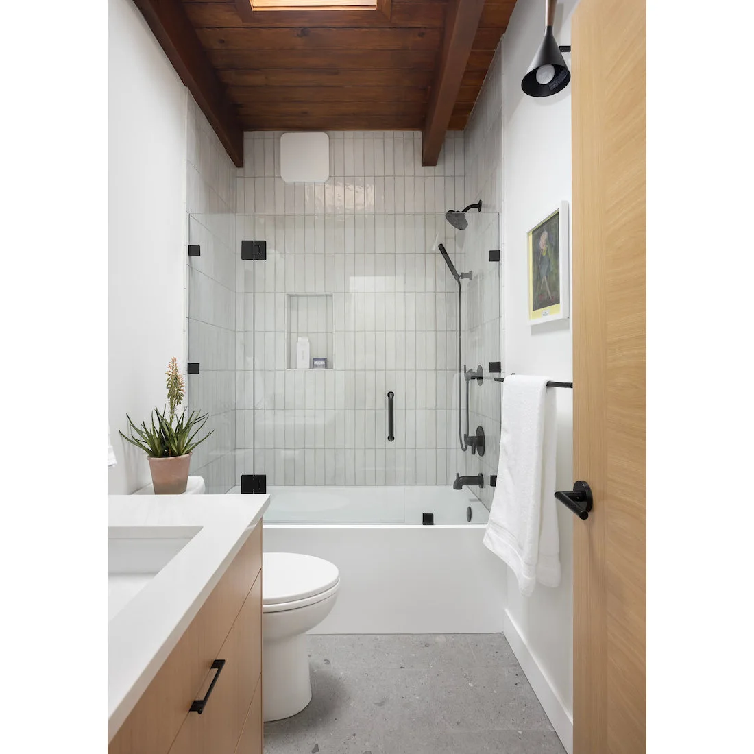 White vertical tiles surround a tub shower beneath a wooden ceiling beside a pale vanity and black fittings