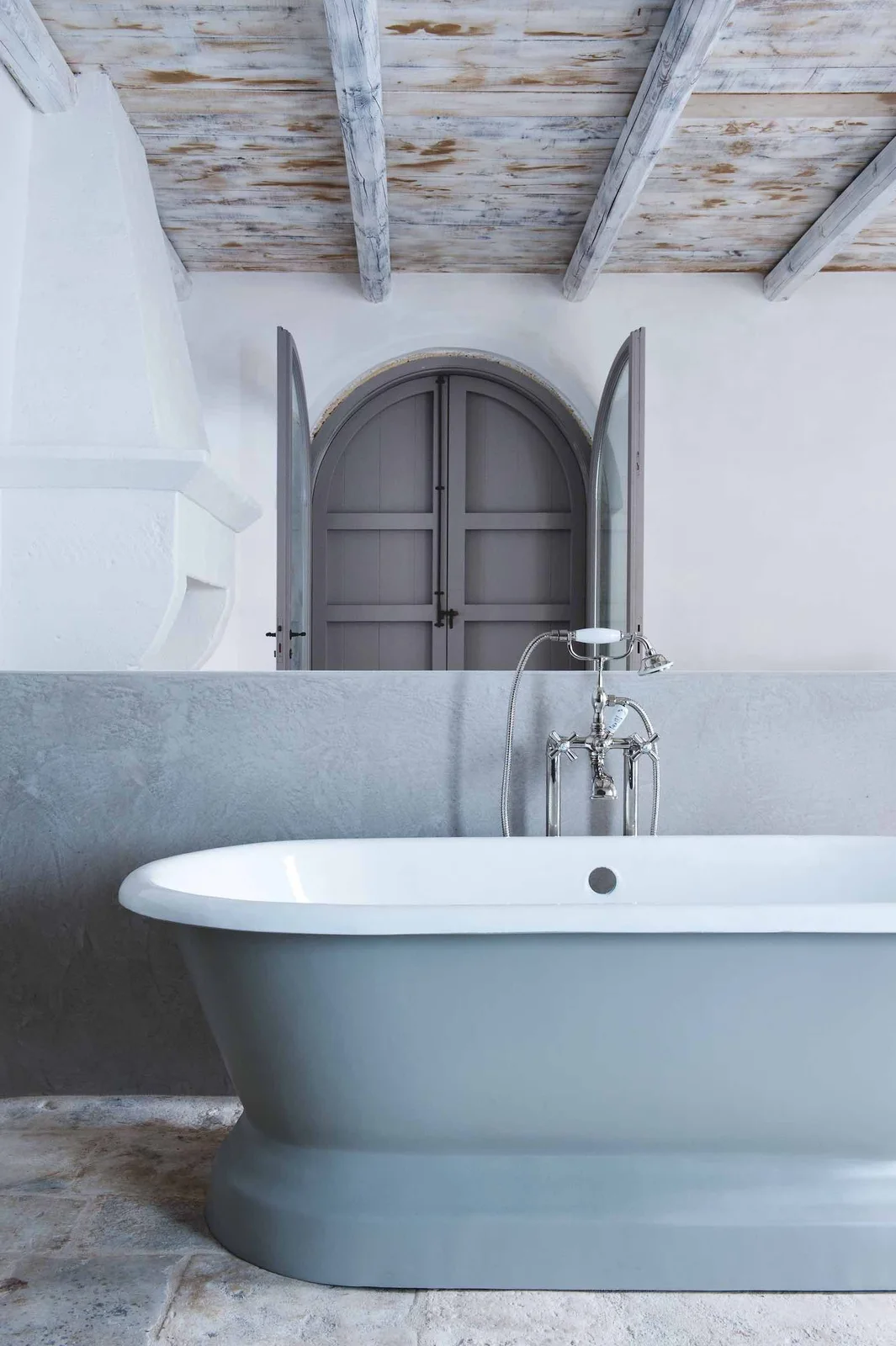 A pale freestanding bath beneath white washed timber boards beside an arched gray door