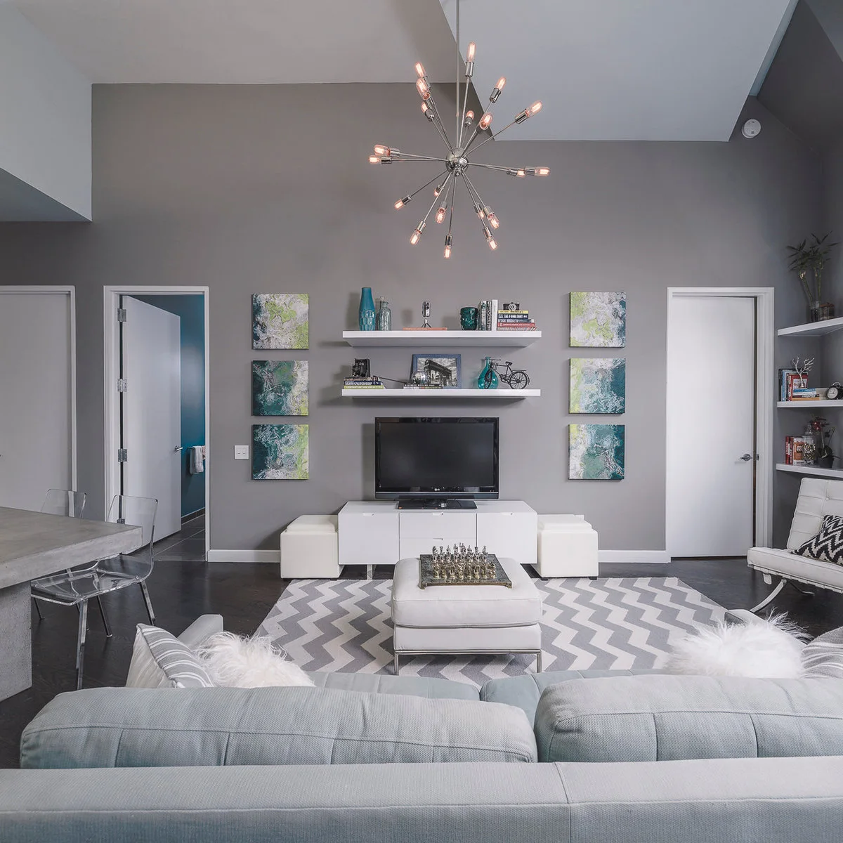 A pale sofa faces a low television unit beneath wall shelves, while transparent dining chairs and a gray table sit at the room’s side.