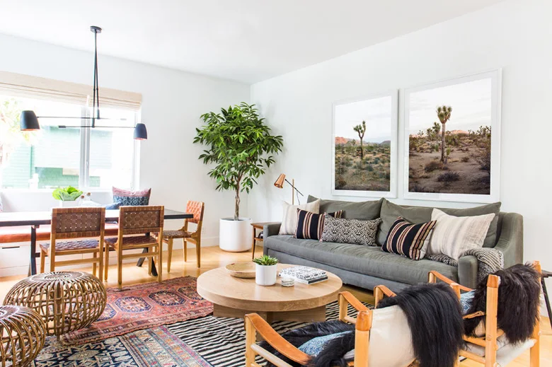 A gray sofa and wood-framed chairs sit around a round wood table on overlapping patterned rugs, with framed desert photographs and woven dining chairs beyond.