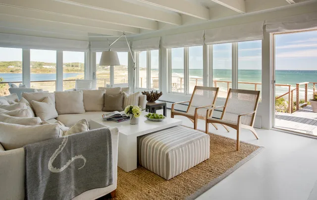 Pale sofas sit beside open timber-framed chairs on a natural-toned rug near generous windows.