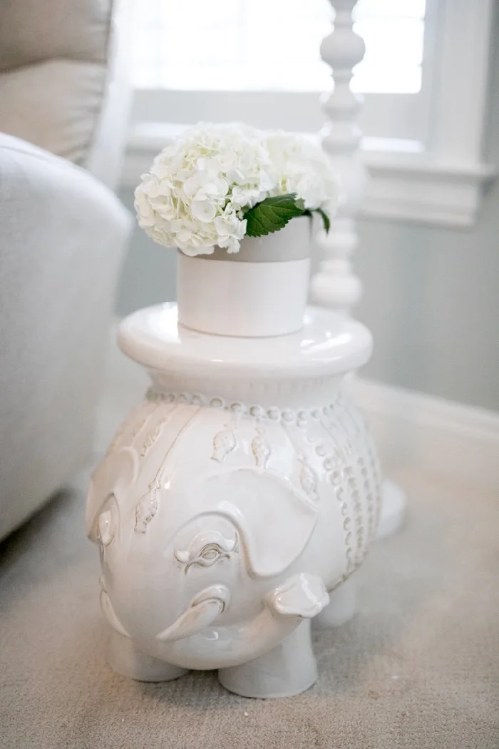 A glossy pale elephant-shaped side table beside a nursery chair holds a pot of white flowers.