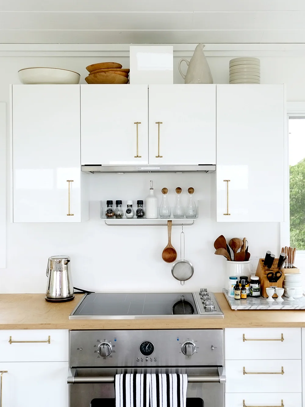 Wood bowls, a pale pitcher, stacked white dishes and a simple frame above glossy white kitchen cabinets with gold-toned handles.