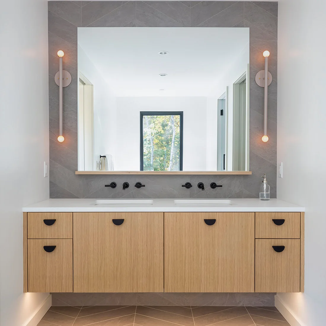 A pale wood double vanity with black half-moon pulls sits beneath a broad mirror, gray veined wall panels and slender vertical globe-ended wall lights.