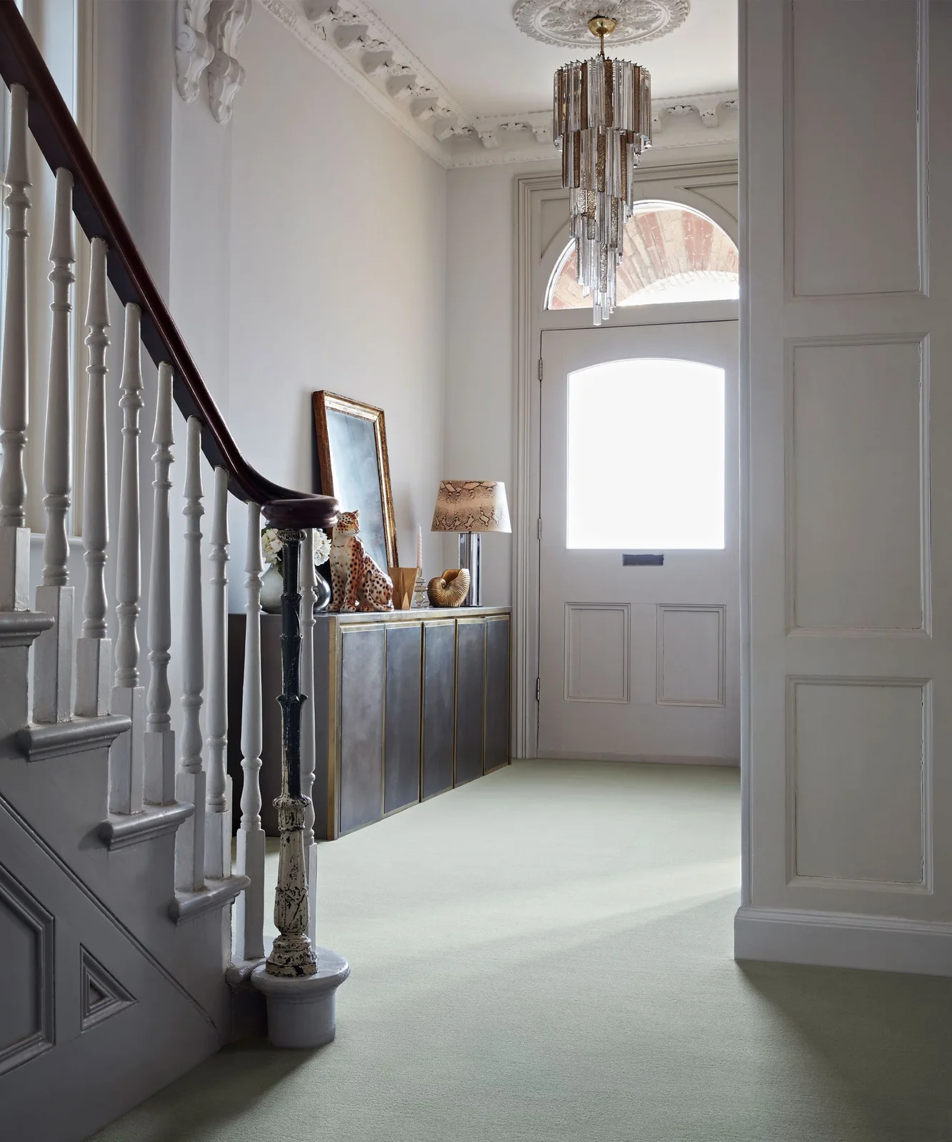A traditional entrance has pale paneled walls and carpet beside a white staircase with a dark handrail, reflective cabinet and hanging clear light
