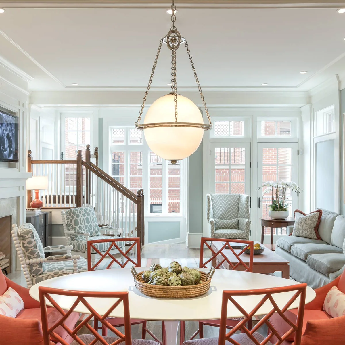 A round white dining table with orange-red chairs shares a pale room with a light sofa, patterned armchairs and a television on the left wall.