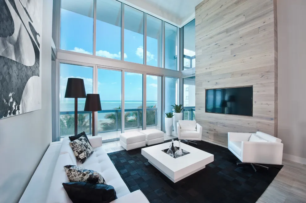A pale plank wall sits behind a television in a room with tall windows and light seating