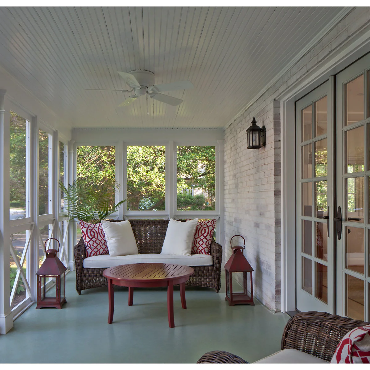 A wicker loveseat with white cushions, red patterned pillows and a red round table on a pale green porch floor.