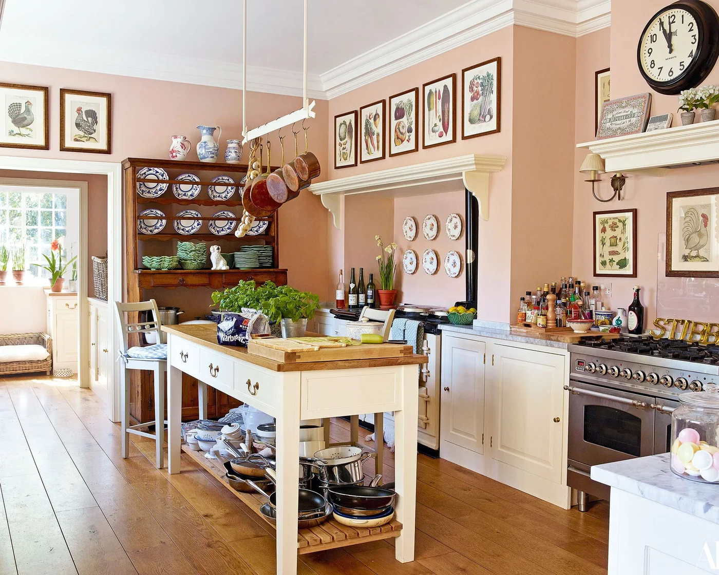 Three patterned pitchers on top of a wood plate dresser in a pink kitchen with blue and white dishes and framed botanical art.