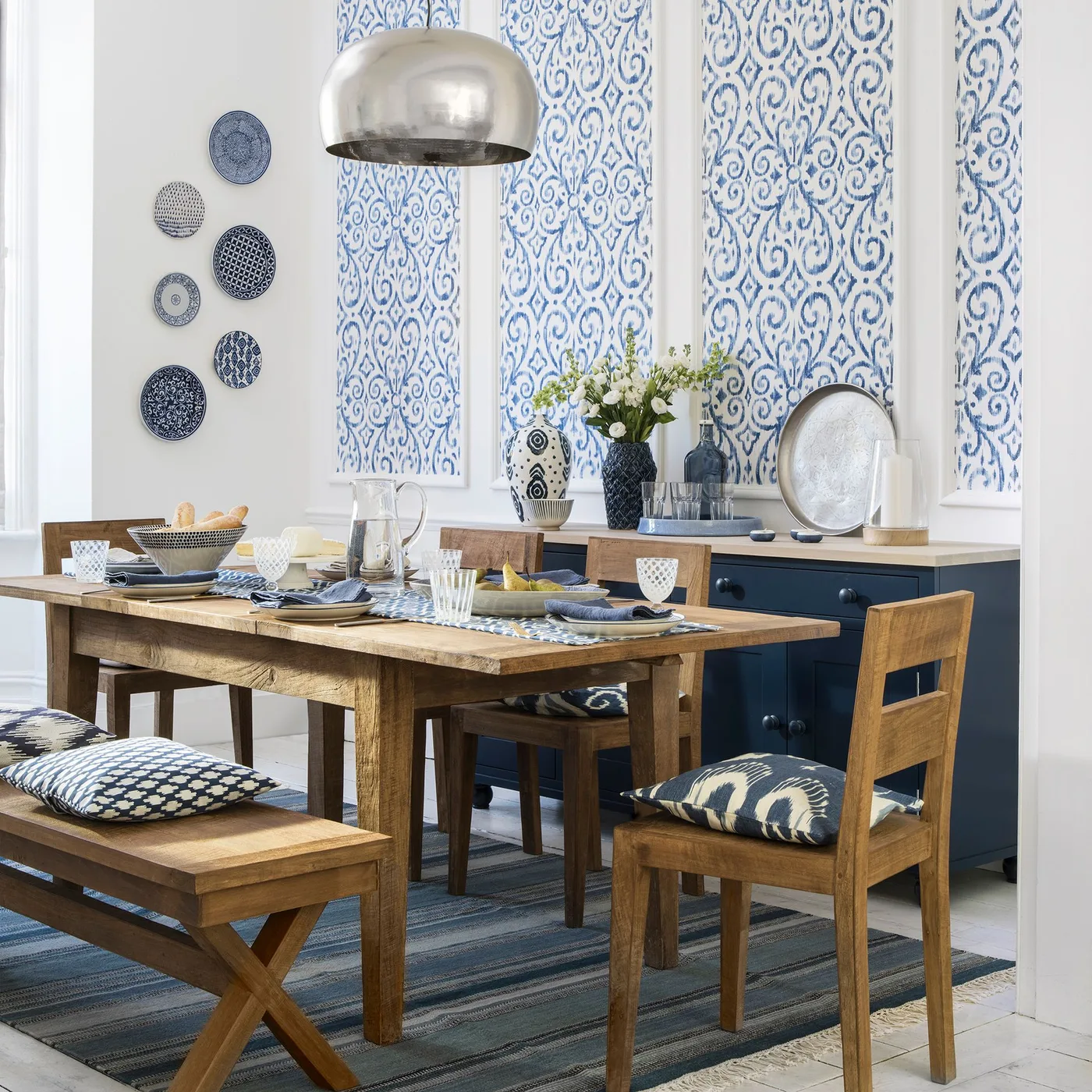 A wood dining table, bench and chairs stand before blue patterned wall panels and a blue sideboard.