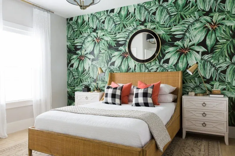 Bedroom with dense green leaf wallpaper, woven bed frame, white bedding, red and checked pillows, pale bedside chests and round mirror.