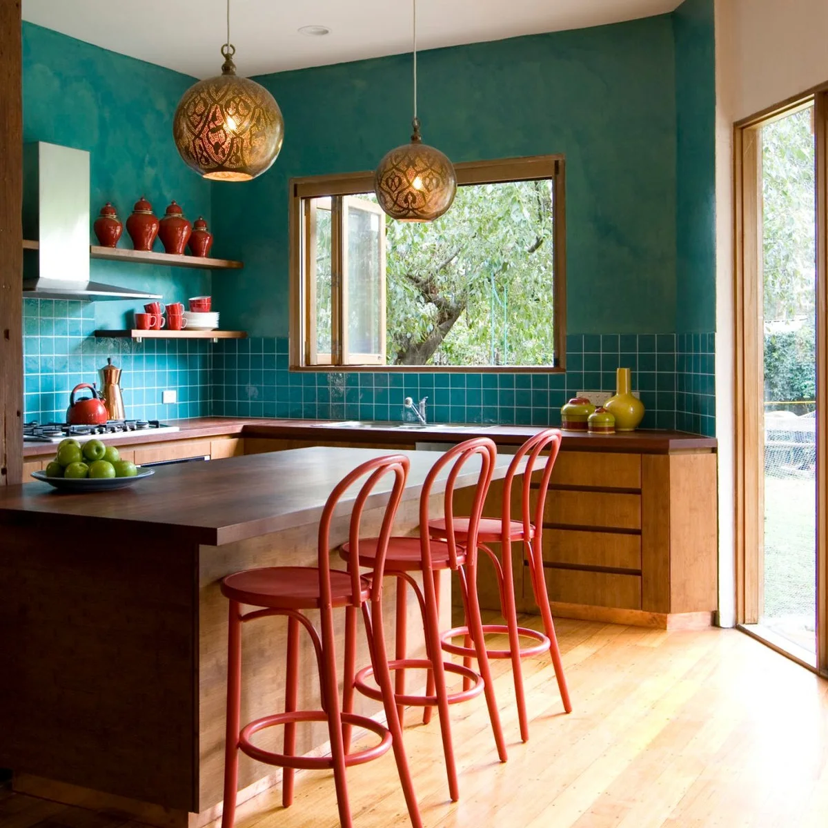 Red cups and lidded vessels on wooden shelves above blue-green tiles in a colorful kitchen.