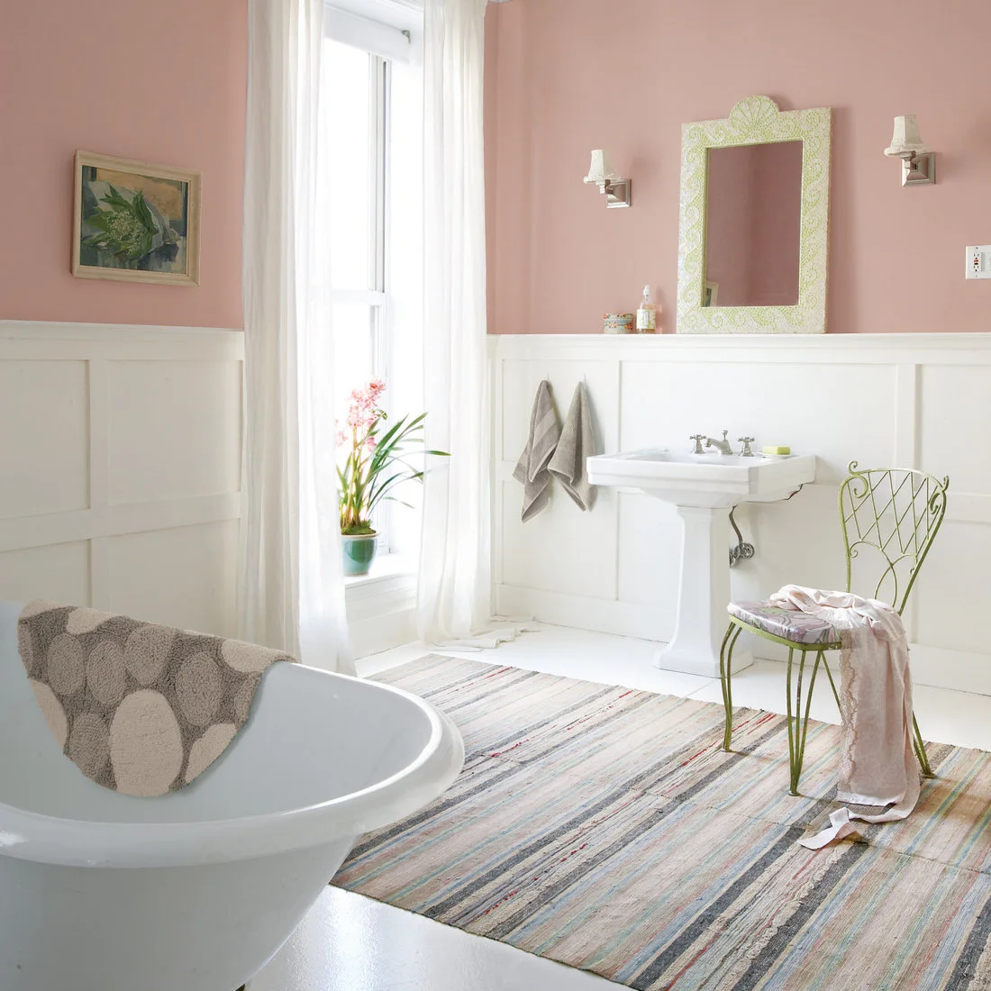 A green patterned mirror frame and slender green chair sit against pink walls above white bathroom paneling.
