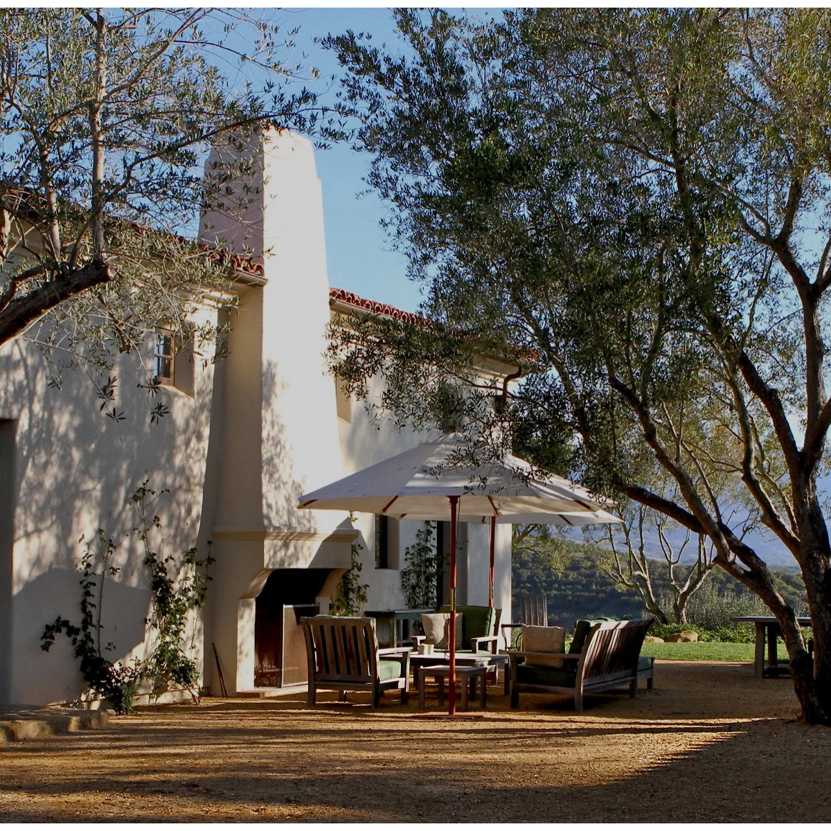 An outdoor sitting area on a fine gravel-like surface beneath trees, with wood chairs, an umbrella and a pale outdoor fireplace.