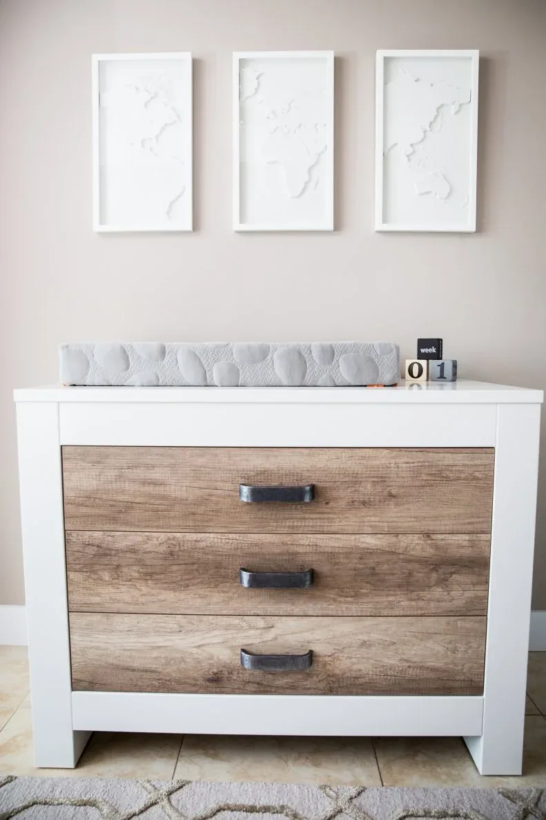 A white changing-area dresser with timber-toned drawer fronts stands beneath three pale framed pictures.