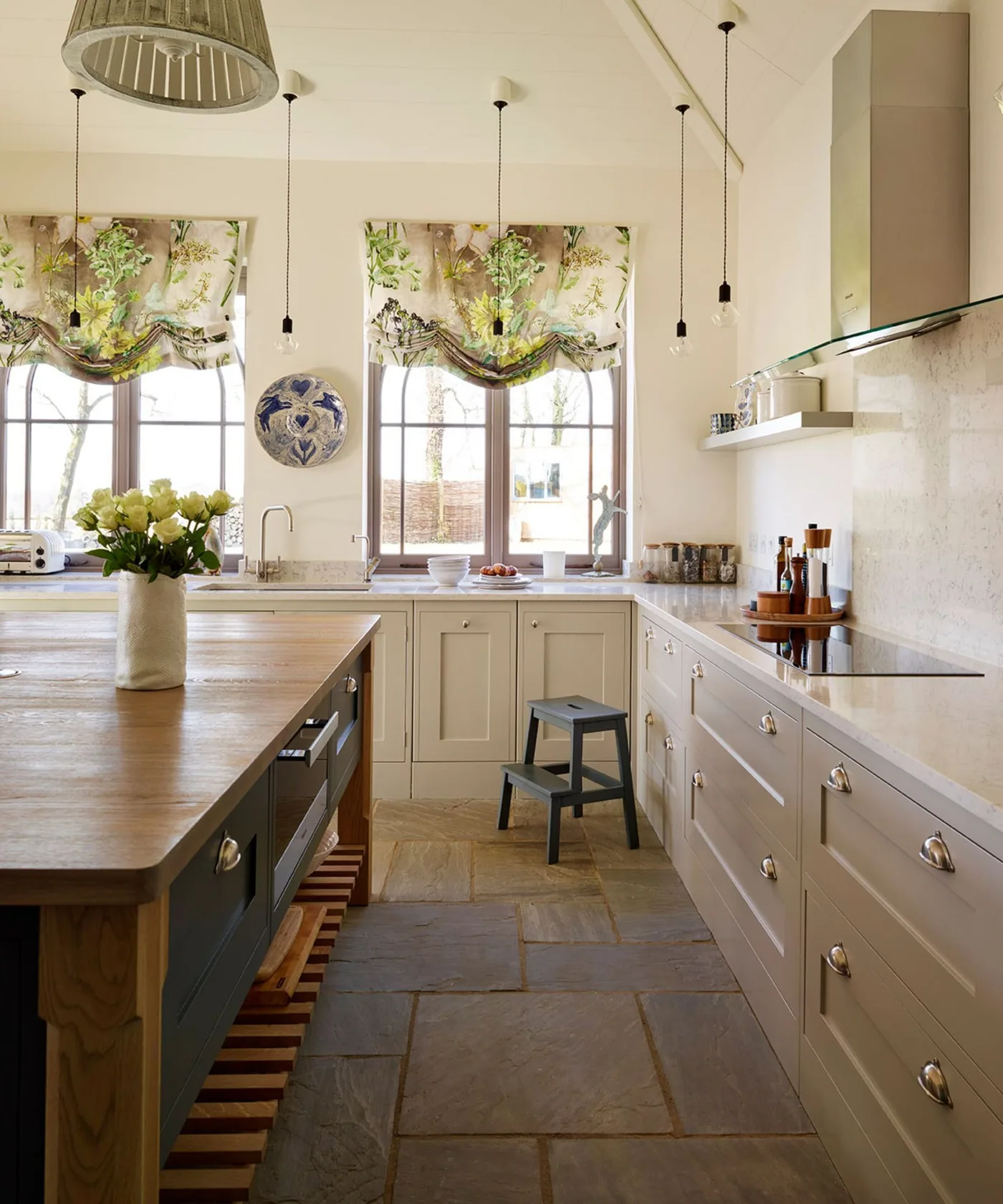 Pale farmhouse kitchen with wooden island, floral window shades, pendant lights and a simple hood.