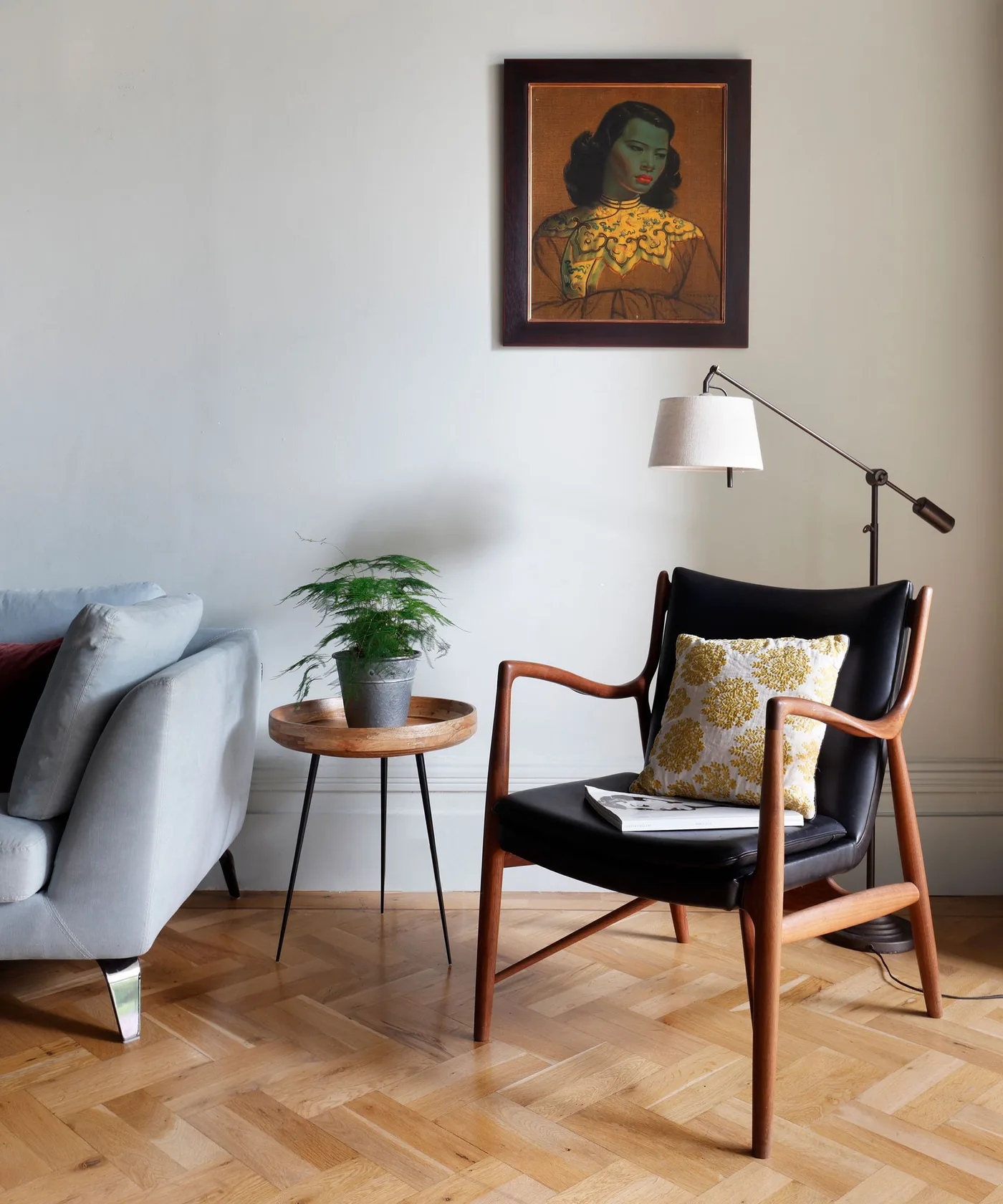 Wood-framed dark reading chair beside a small round wood table, pale wall and warm patterned parquet floor