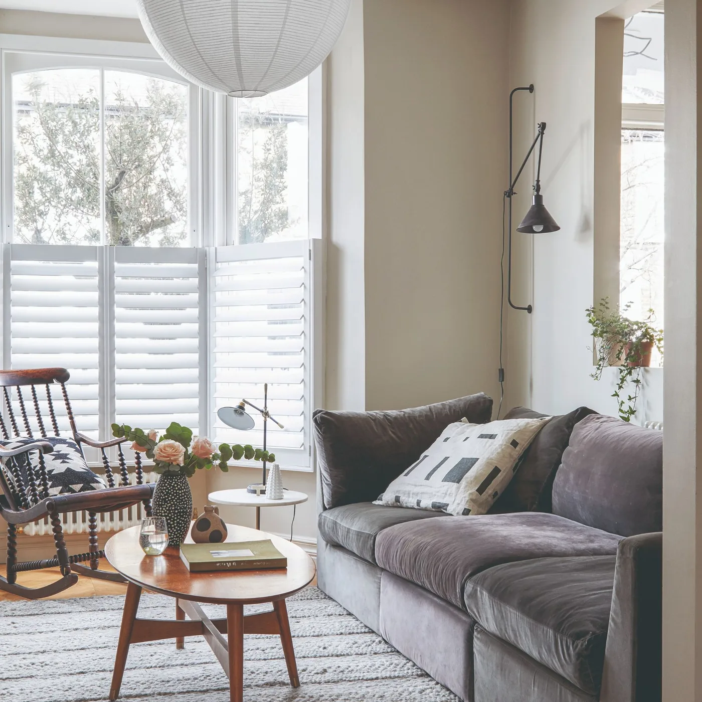 Gray sofa beside a round wood coffee table, pale shutters, a wood rocking chair and an adjustable wall lamp