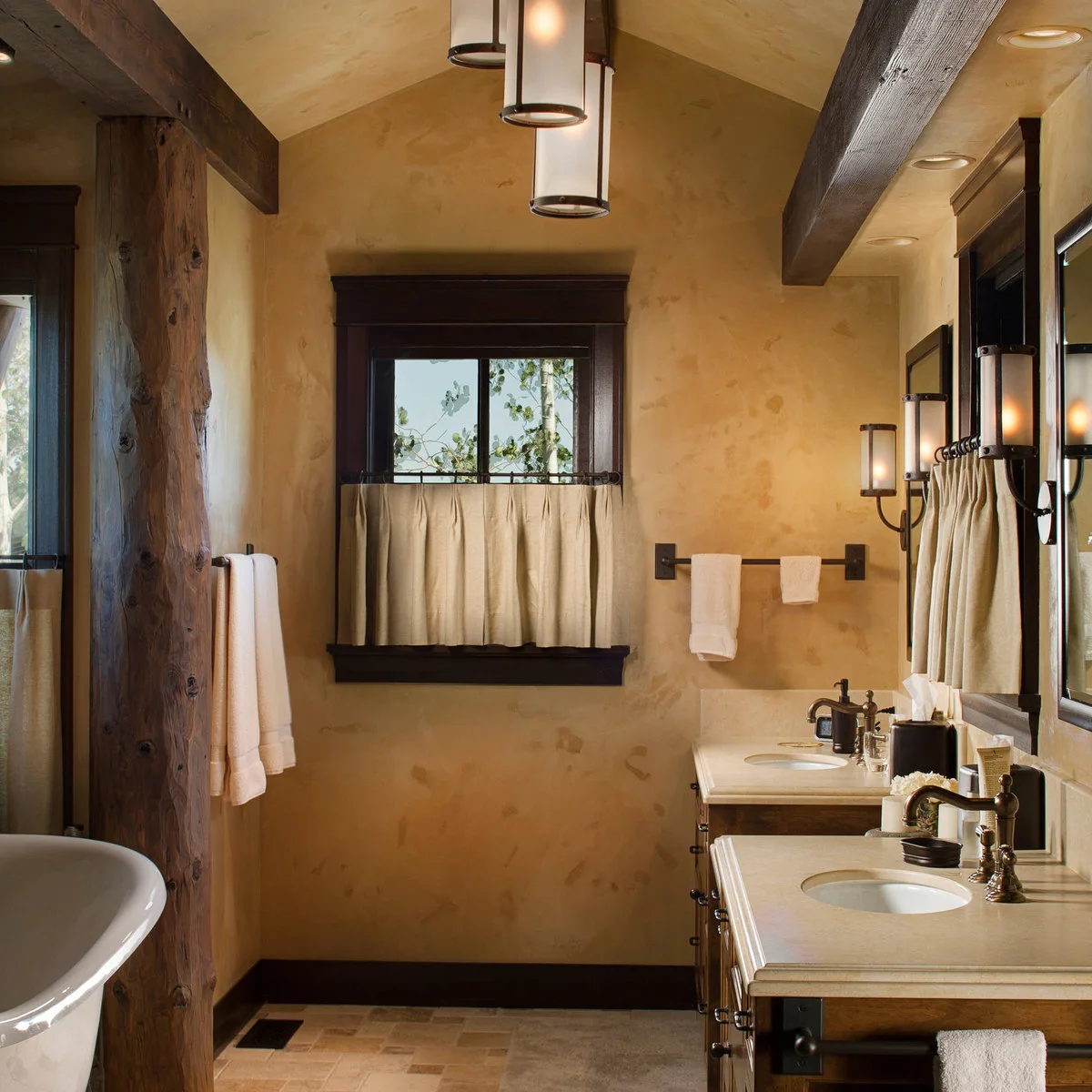 Plain cream cafe curtains cover the lower half of a dark-framed window in a warm-toned bathroom with exposed timber.