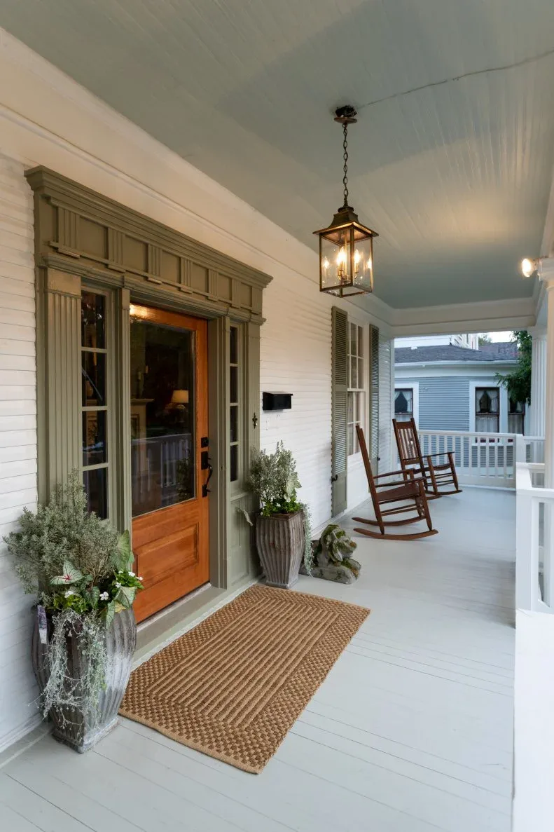 Warm wood glazed door with detailed surround, pale porch boards, lantern, dark mat and wood rocker