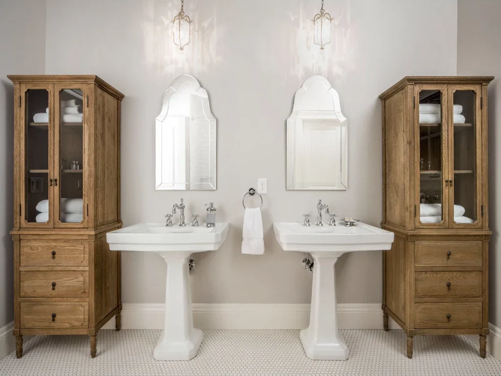 Two white pedestal sinks sit between tall wood storage cabinets beneath shaped mirrors and small hanging lights.