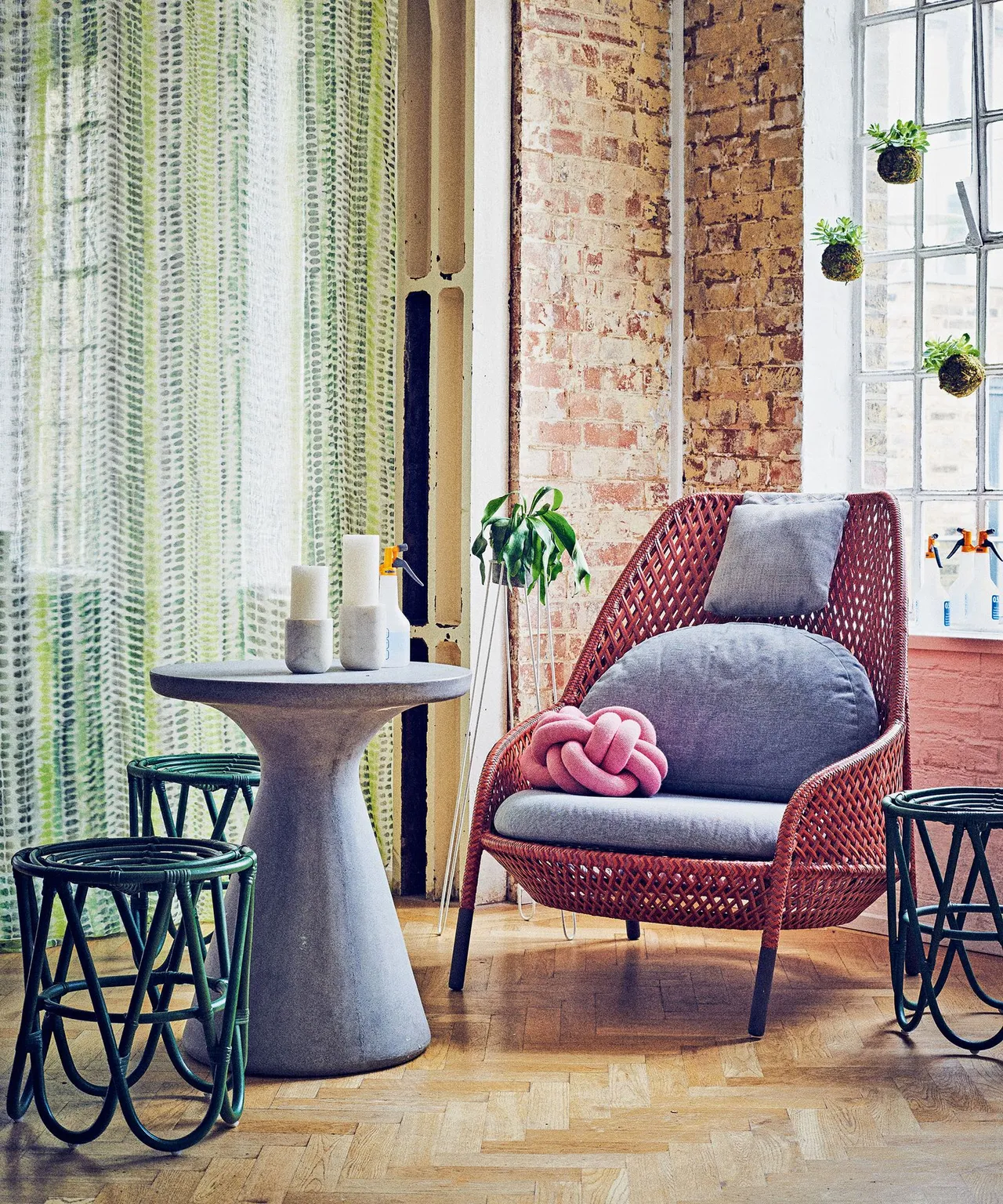 A red woven chair with gray cushions sits beside a pale pedestal table, patterned curtains and a brick wall.