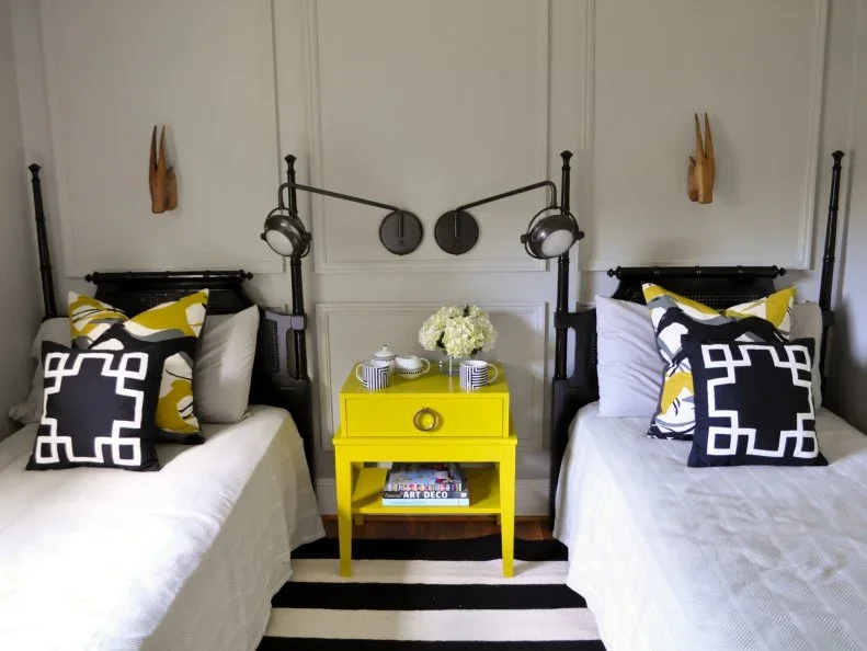 Two black metal twin beds have graphic black-and-white cushions beside a bright yellow shared table beneath adjustable black wall lights.