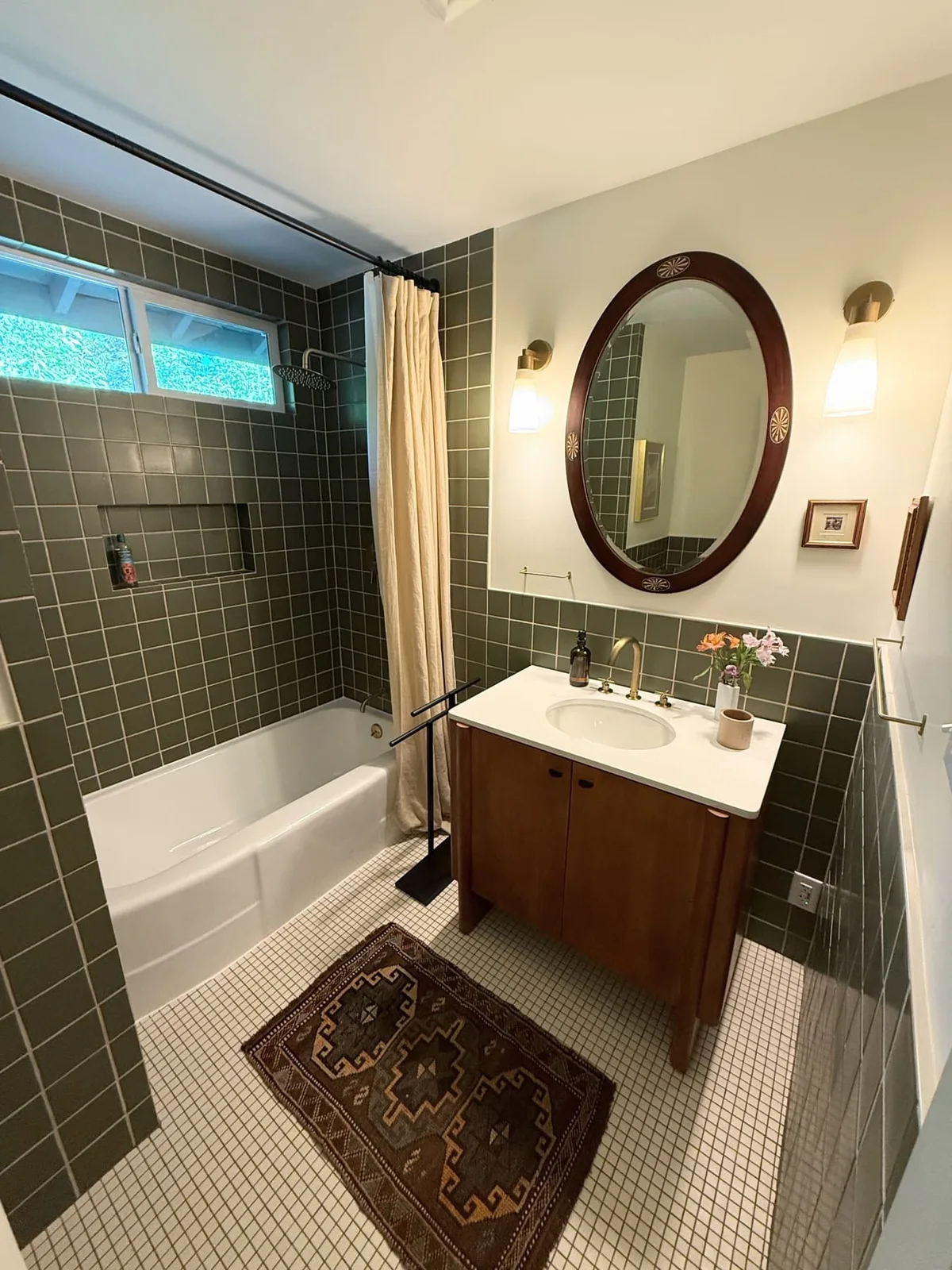 Olive square tiles line a compact tub-shower and continue behind a wood vanity with an oval mirror and cream upper walls.