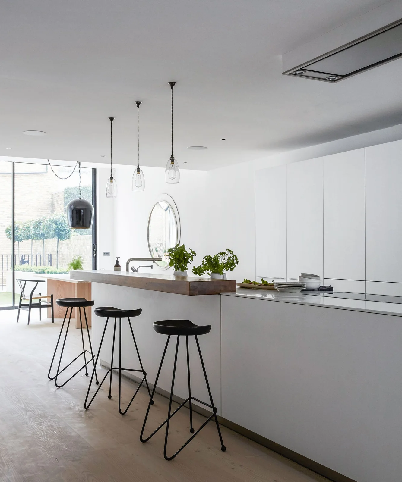 A raised wooden ledge sits on a pale kitchen island beside black stools and white cabinets