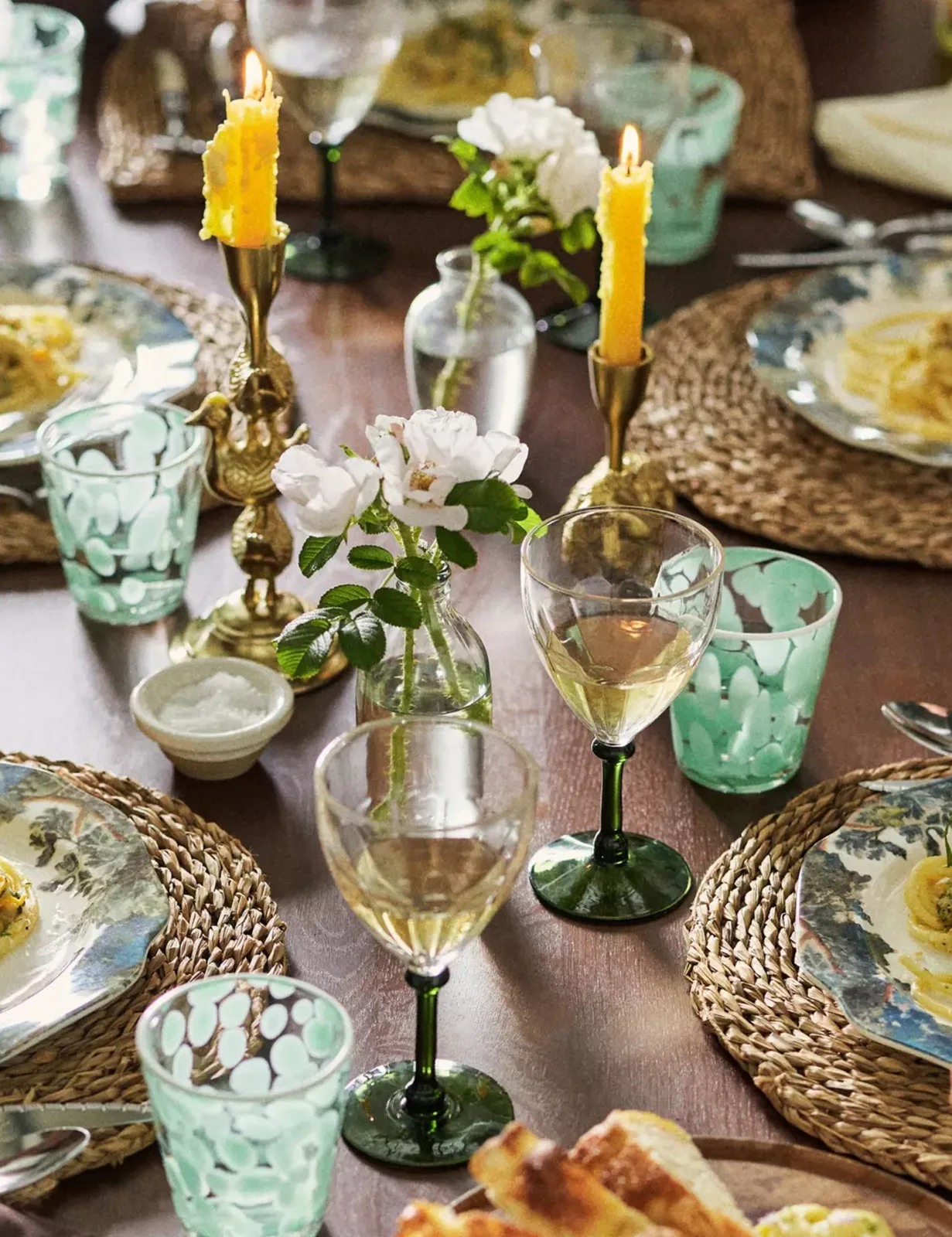 Green-stemmed wine glasses and pale green tumblers sit beside floral plates, woven chargers and small clear flower vases