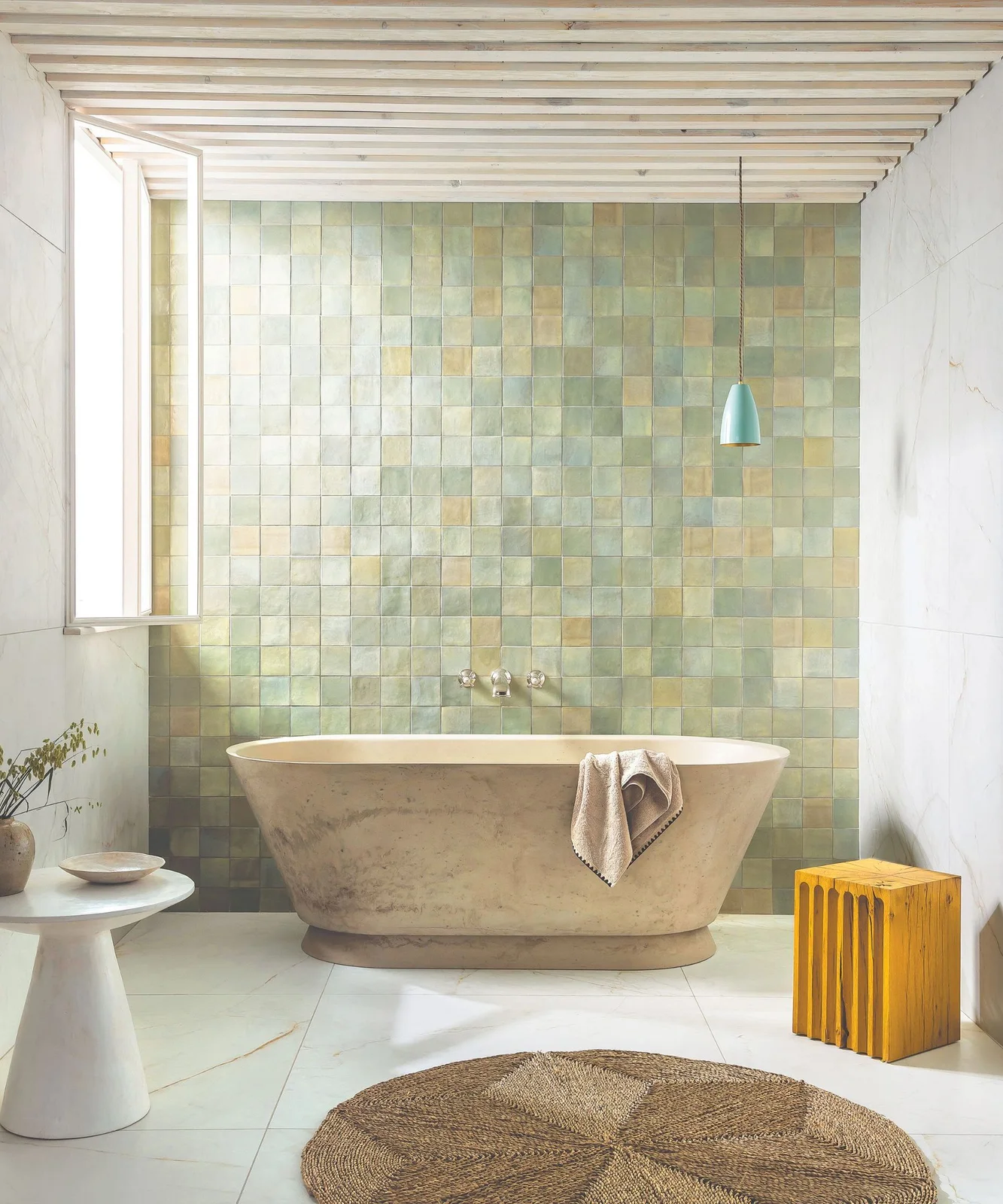 A pale rounded bath stands before muted green and yellow square tiles beneath pale ceiling slats beside a yellow stool and white table