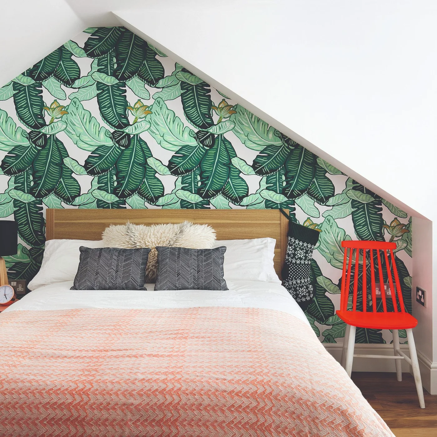 Large green leaf wallpaper fills a sloped bedroom wall behind a wood headboard, white bedding, peach cover and bright red chair