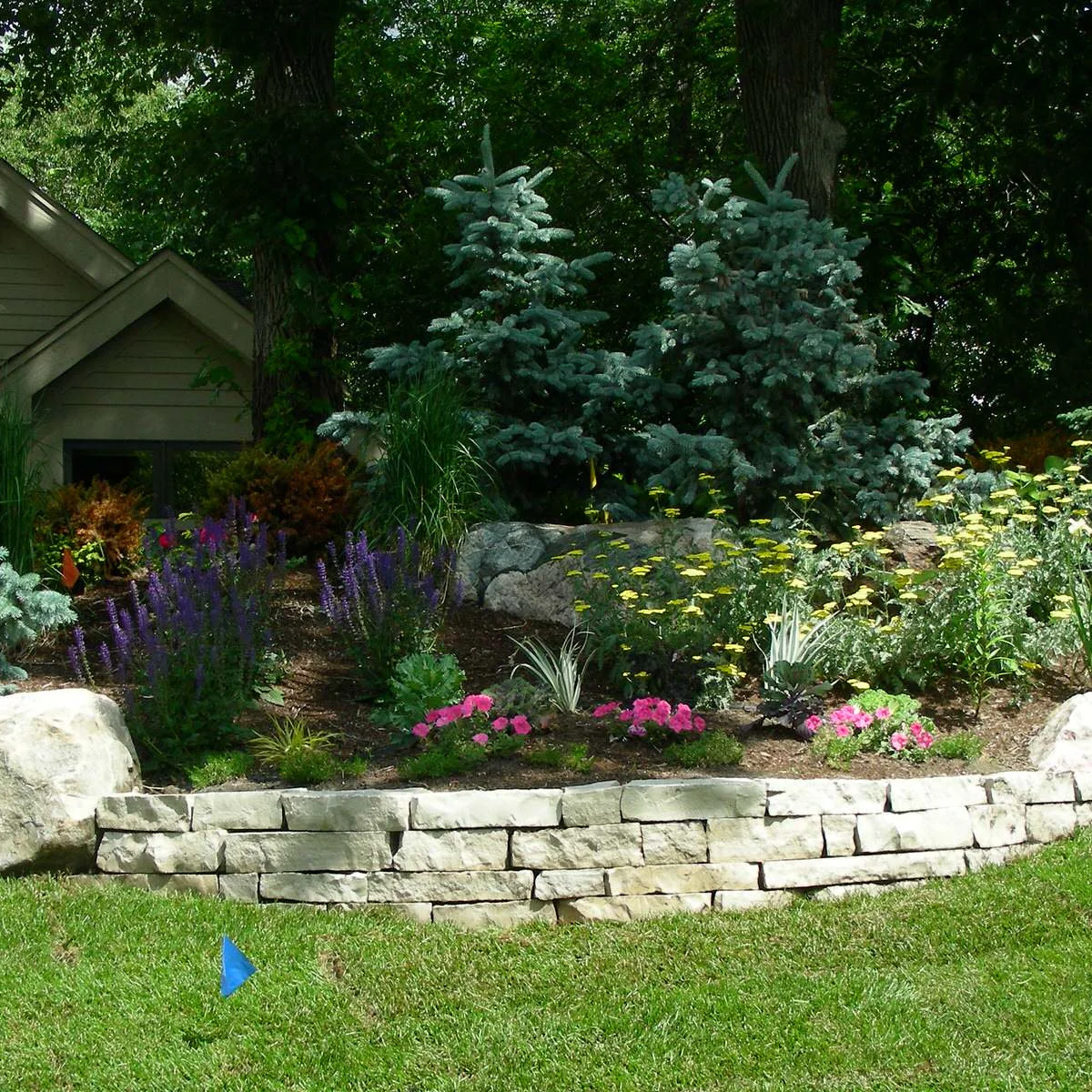 A low pale block retaining wall supports a sloping flower border with boulders near its ends.