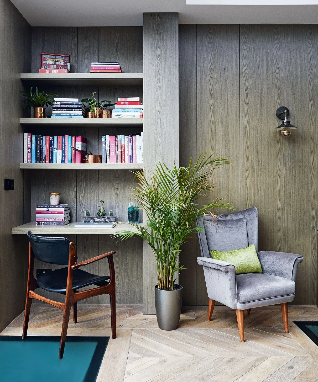 A narrow desk alcove with shelves sits within a wood-grain wall beside a gray armchair, a plant and a black wall light.