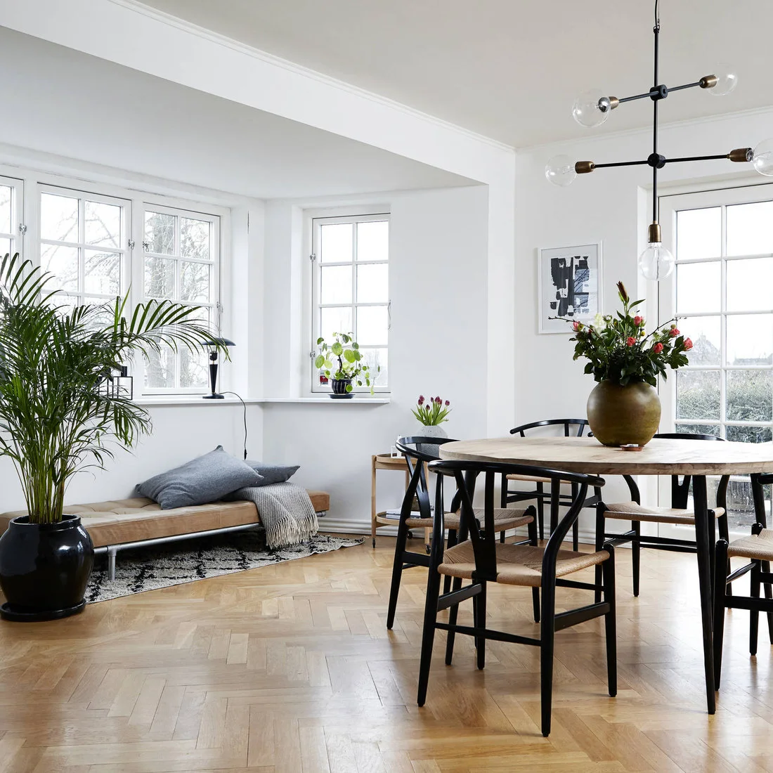 A round wood dining table and black woven-seat chairs stand beside white walls, broad windows and a low cushioned bench.