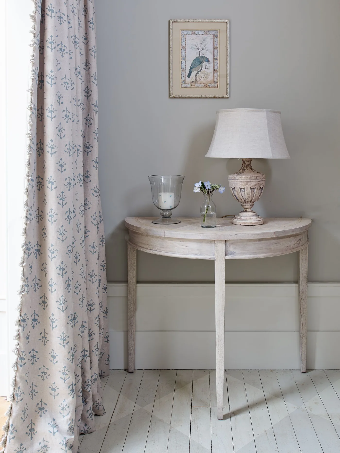 Pale entry corner with a half-round console table, shaded lamp, framed bird picture and a light patterned curtain.