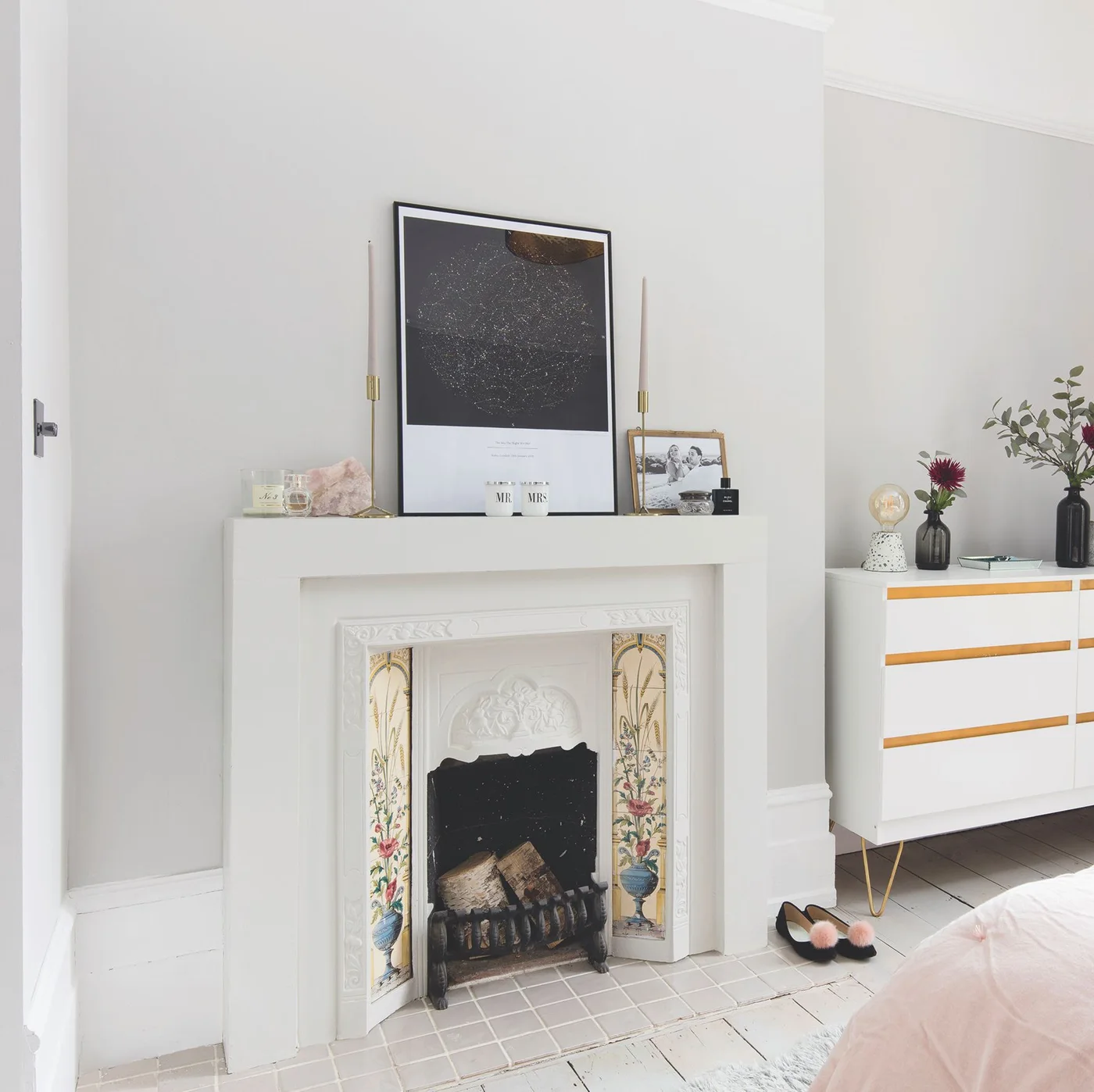 Slender floral tile panels flank a bedroom fireplace beneath a white mantel, with a pale tiled hearth and light gray walls.