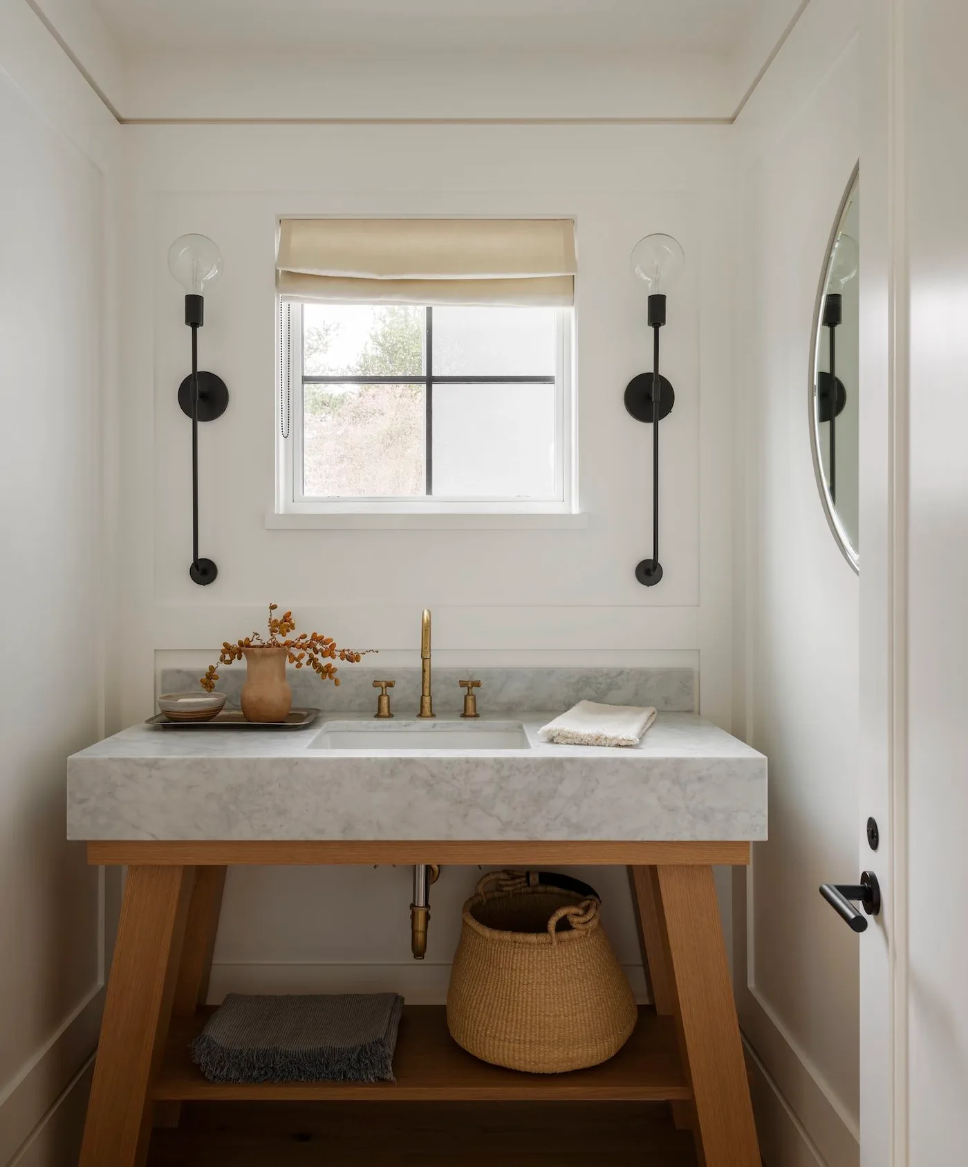 White powder room with pale veined basin on wood trestle-style washstand, black lights and lower basket