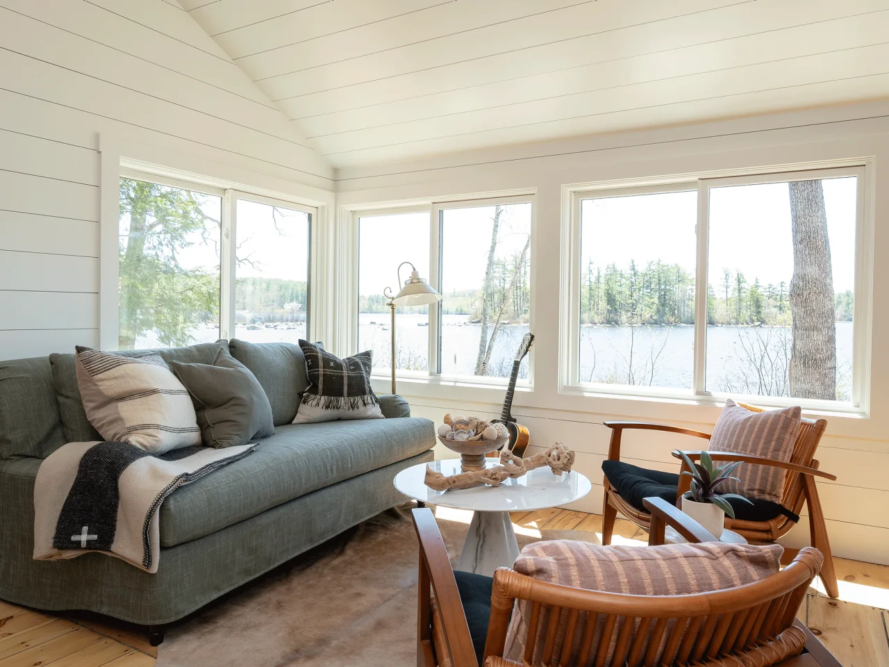 A muted gray-green sofa and timber chairs sit around a round white table in a pale paneled room overlooking water.
