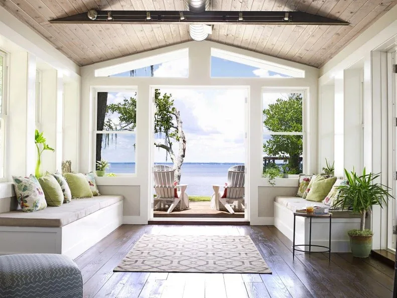 Bright sunroom with built-in benches, pale cushions, a patterned runner and doors opening toward the water.